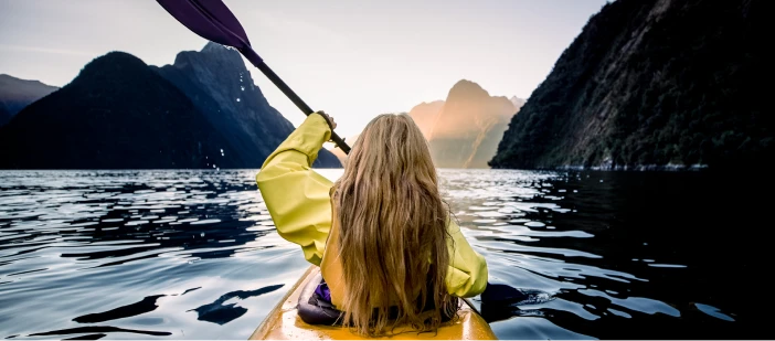 A woman in a kayak on the water in New Zealand, with beautiful mountains in the distance.