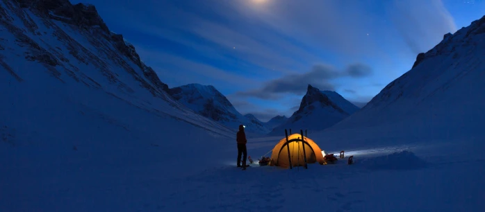 A person standing next to an illuminated tent in a snowy valley.