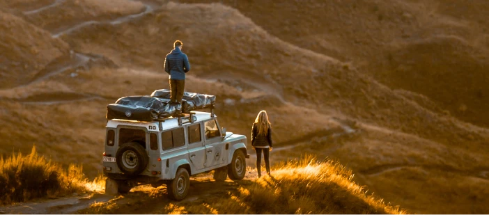 A man and a woman with a Land Rover overlooking hills in New Zealand.