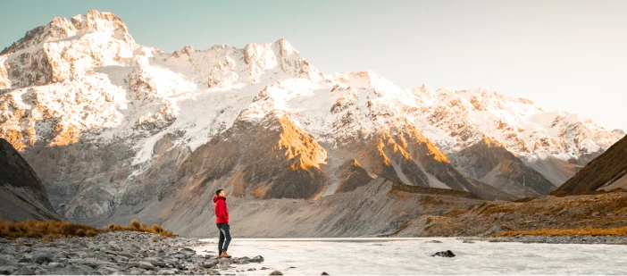 A person standing next to a river with mountains in the background in New Zealand.