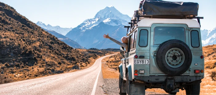 A person waving from the window of a Land Rover with New Zealand mountains in the background.